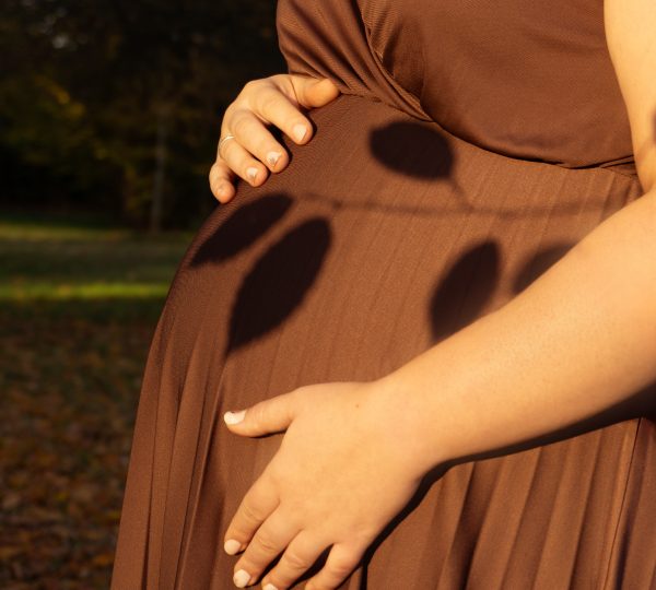 Detailaufnahme eines Babybauchs beim Outdoor-Schwangerschaftsshooting: werdende Mama im braunen Kleid hält den Bauch im warmen Abendlicht – minimi fotografie.