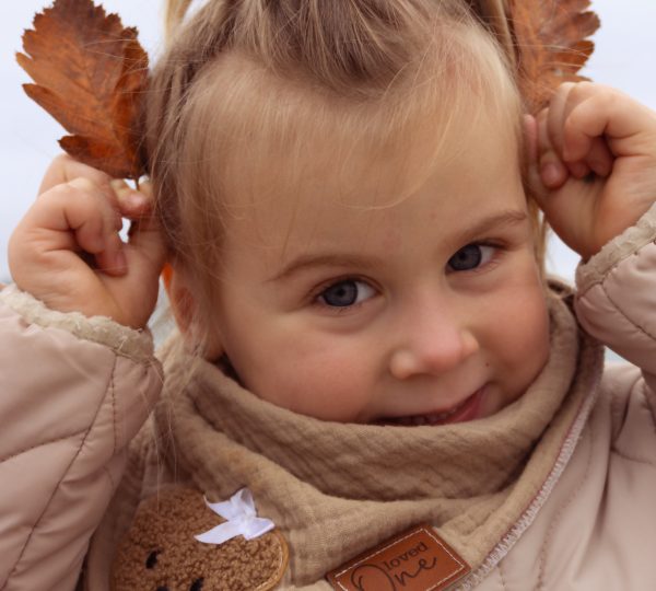 Nahaufnahme eines Kindes am Strand: Mädchen mit Schal und Jacke hält Herbstblätter an die Ohren und lächelt – natürliche Kinderfotografie outdoor, minimi fotografie.