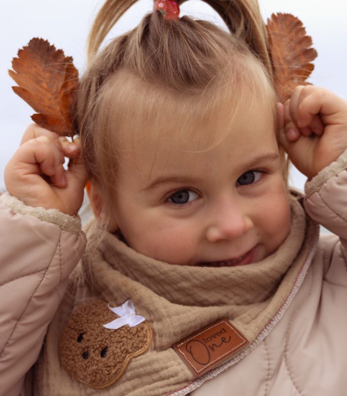 Nahaufnahme eines Kindes am Strand: Mädchen mit Schal und Jacke hält Herbstblätter an die Ohren und lächelt – natürliche Kinderfotografie outdoor, minimi fotografie.